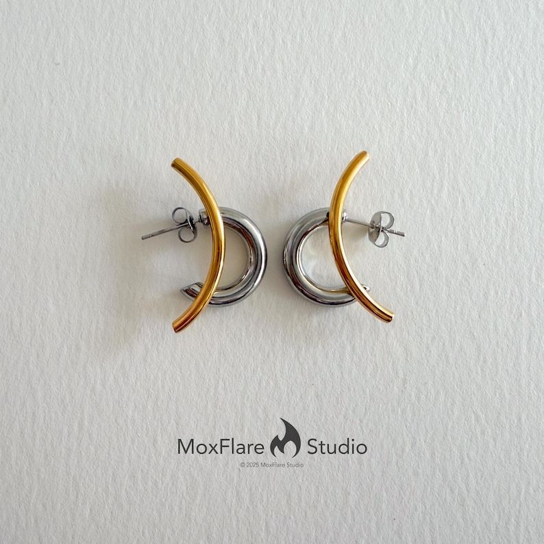 May include: A pair of modern earrings with a gold-colored curved bar accenting a silver-colored hoop. The earrings are displayed on a white surface. The brand name "MoxFlare Studio" is printed in black text.