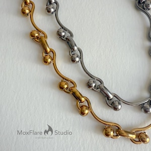 May include: Close-up of two necklaces. One is gold-toned, the other is silver-toned. Both feature a chain design with small, round beads. The gold necklace is on the left, and the silver necklace is on the right. The MoxFlare Studio logo is in the bottom left corner.