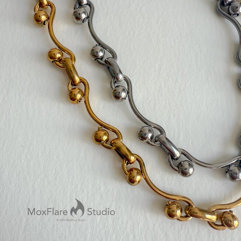 May include: Close-up of two necklaces. One is gold-toned, the other is silver-toned. Both feature a chain design with small, round beads. The gold necklace is on the left, and the silver necklace is on the right. The MoxFlare Studio logo is in the bottom left corner.