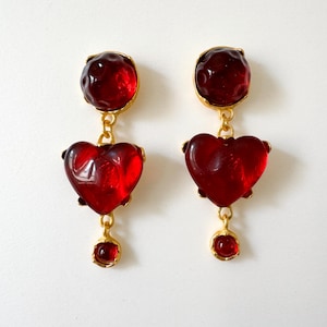 May include: A pair of red glass heart-shaped dangle earrings with gold accents. The earrings feature a round red glass bead at the top and a smaller red glass bead at the bottom.
