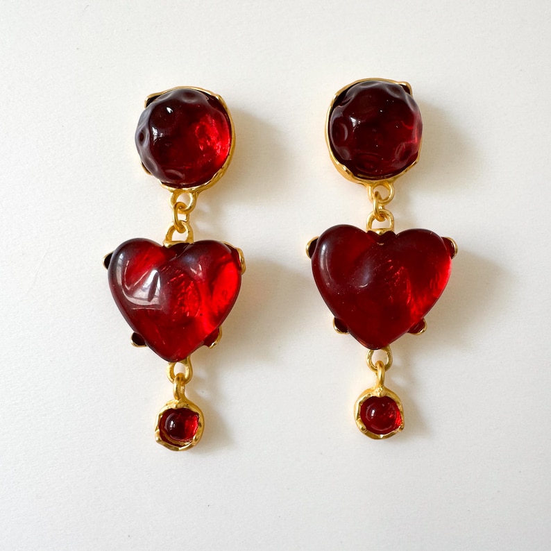 May include: A pair of red glass heart-shaped dangle earrings with gold accents. The earrings feature a round red glass bead at the top and a smaller red glass bead at the bottom.