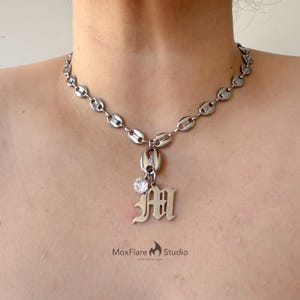 以下が含まれることがあります： 楕円形のリンクのチェーンが付いたシルバーカラーのネックレス。装飾的なフォントの文字「M」と小さく丸い透明な宝石が付いたペンダントがチェーンからぶら下がっています。ネックレスは人の胸に表示されています。