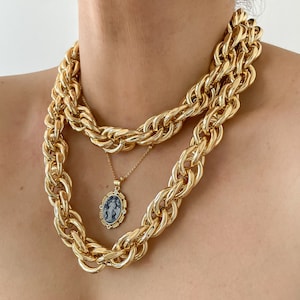 May include: A gold chain necklace with a thick rope chain design. The necklace is layered with a thinner chain with a pendant featuring a cameo of a woman in a blue and white design.