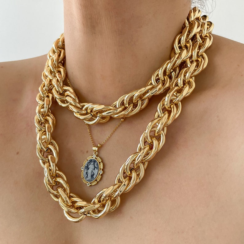 May include: A gold chain necklace with a thick rope chain design. The necklace is layered with a thinner chain with a pendant featuring a cameo of a woman in a blue and white design.