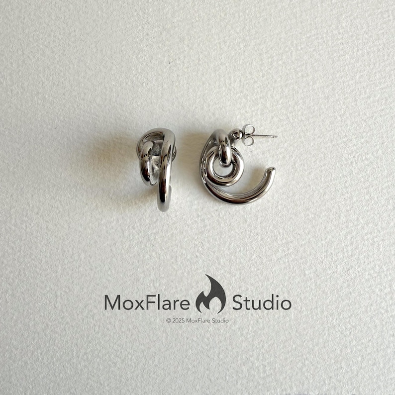 May include: A pair of silver-toned hoop earrings with a unique knot design. The earrings have a curved, open-ended shape and a post closure. The earrings are displayed on a white surface. The MoxFlare Studio logo is visible.
