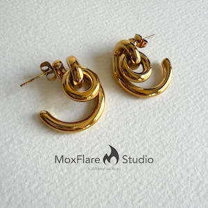 May include: A pair of gold-colored earrings with a unique, looped design. Each earring features a curved, open-ended shape connected to a circular element, creating a stylish and modern look. The earrings are displayed on a white surface.