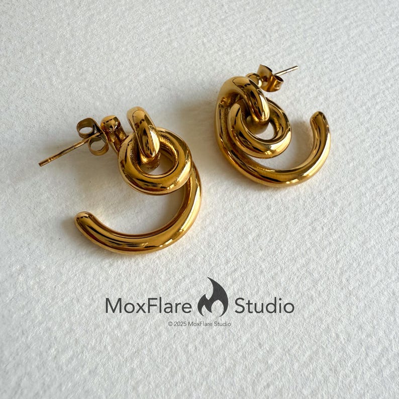 May include: A pair of gold-colored earrings with a unique, looped design. Each earring features a curved, open-ended shape connected to a circular element, creating a stylish and modern look. The earrings are displayed on a white surface.