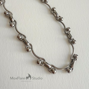 May include: A silver-toned necklace with a unique design. The necklace features a series of curved, wavy links connected by small, round beads. The MoxFlare Studio logo is visible in the bottom left corner.