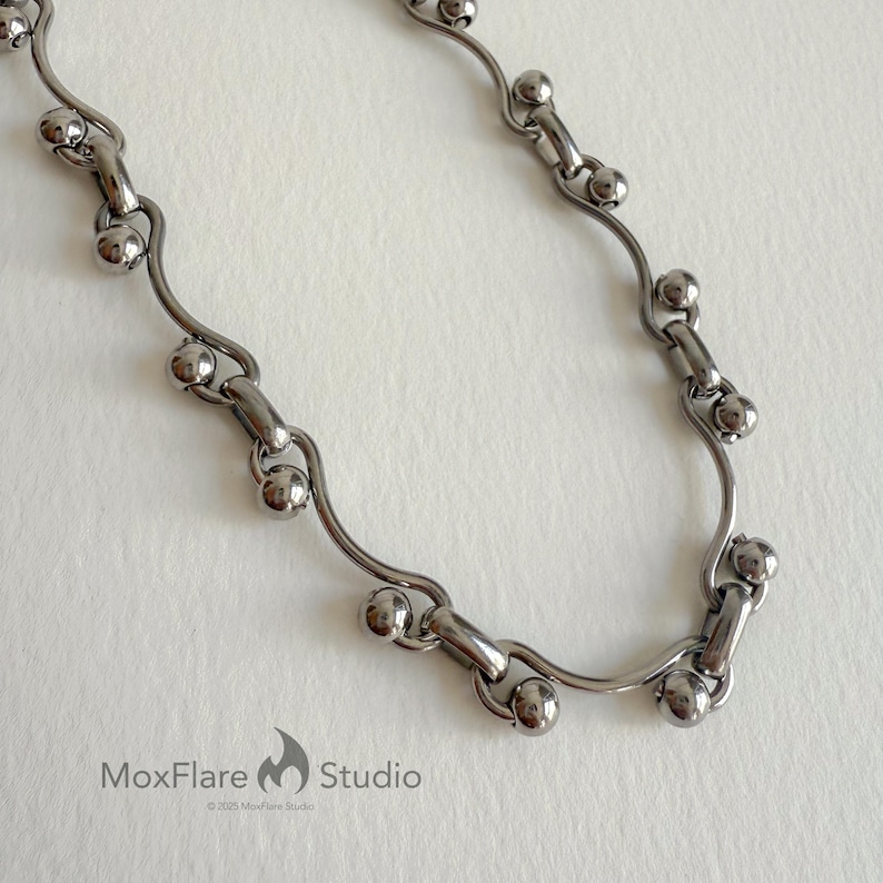 May include: A silver-toned necklace with a unique design. The necklace features a series of curved, wavy links connected by small, round beads. The MoxFlare Studio logo is visible in the bottom left corner.