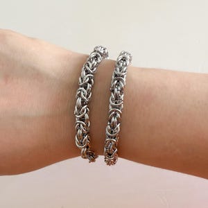 Pu&ograve; includere: Due braccialetti a catena argentati sul polso di una persona. I braccialetti sono fatti di anelli di metallo intrecciati.
