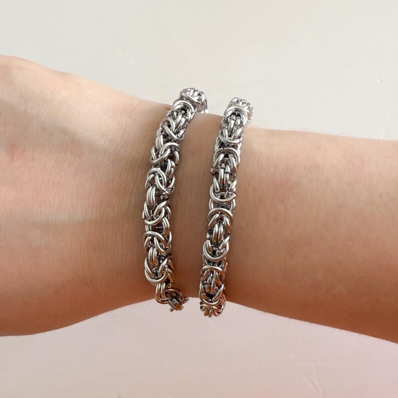Pu&ograve; includere: Due braccialetti a catena argentati sul polso di una persona. I braccialetti sono fatti di anelli di metallo intrecciati.
