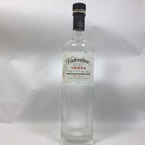 May include: A clear glass Valentine vodka bottle with a black cap and a white label. The label features the brand name, product details, and the text "Detroit". The bottle has vertical ridges on the lower half.