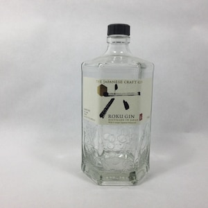 May include: A clear, hexagonal glass bottle with a black cap. The label features the text 'THE JAPANESE CRAFT GIN', 'ROKU GIN', and 'DISTILLED IN JAPAN' with black brushstroke graphics. The lower portion of the glass bottle has embossed floral patterns.