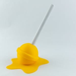 May include: A whimsical, yellow lollipop sculpture with a white stick, appearing to melt onto a matching yellow surface. The lollipop is a bright, solid color, creating a playful and eye-catching design.