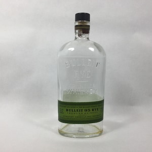 May include: A clear glass Bulleit 95 Rye Frontier Whiskey bottle with a black cap. The bottle has a green label with white text. The bottle is empty and appears to be a standard size.