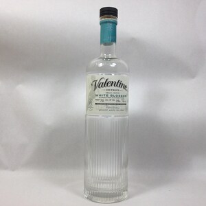 May include: A clear glass bottle of Valentine Detroit White Blossom gin. The bottle has a black cap, a teal neck wrap, and a label with the brand name and product details. The bottle has vertical ridges.