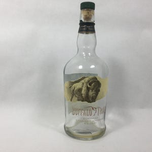 May include: An empty clear glass Buffalo Trace Kentucky Straight Bourbon Whiskey bottle with a dark green cap and a cork stopper. The front label features a tan background with a brown illustration of a buffalo and the brand name text.