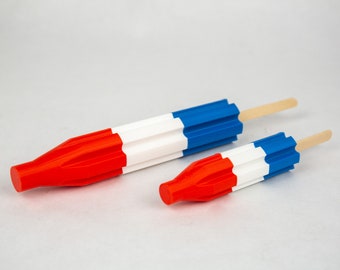 May include: Two red, white, and blue popsicle-shaped objects with wooden sticks. The larger one is approximately 6 inches long, and the smaller one is about 3 inches long. The popsicles are a patriotic design.