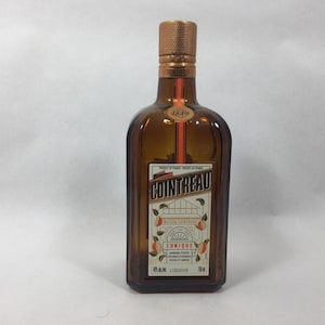 May include: A rectangular amber glass Cointreau liqueur bottle with a textured copper-colored cap. The front label features the brand name, the year 1849, orange illustrations, and the text L'UNIQUE. The bottle has a volume of 750 ml.