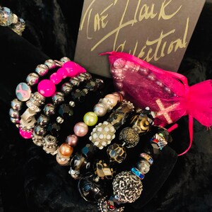 May include: A stack of colorful beaded bracelets with pink, black, and silver beads. The bracelets are arranged on a black fabric background. The text "The Flair Collection" is visible on a gray box in the background.