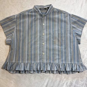 May include: A short-sleeved button-down shirt featuring a vertical striped pattern in shades of blue, white, and tan. The shirt has a mandarin collar, a single chest pocket on the left side, and a gathered ruffle hem along the bottom edge.