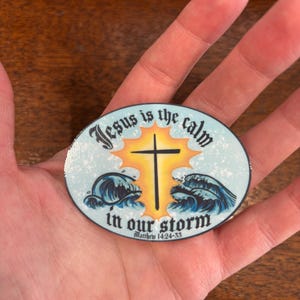 May include: A round sticker with a blue, white, and yellow design. The sticker features a cross with a sun behind it and waves in front of it. The text on the sticker reads "Jesus is the calm in our storm" and "Matthew 14:24-33".