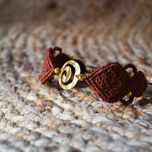 Op de afbeelding: Een bruin macramé armband met een gouden spiraalbedel en gouden kralen.