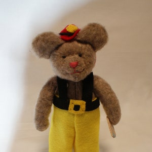 May include: A handmade brown teddy bear wearing a firefighter's hat and uniform. The bear has a red nose, yellow pants, a black belt with a gold buckle, and a small wooden tool. The hat is red with yellow accents.