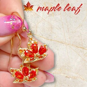 May include: Gold dangle earrings featuring a maple leaf design. The leaves are decorated with red and orange gemstones.