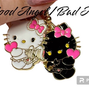 May include: A pair of gold-tone charm earrings featuring a white Hello Kitty with pink accents and a black Hello Kitty with pink accents. The white Hello Kitty is holding a pink heart and has angel wings. The black Hello Kitty is holding a pink heart and has devil wings. The earrings are labelled "Good Angel/Bad Angel".