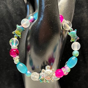 May include: A colourful beaded bracelet with silver turtle charms. The bracelet features pink, blue and clear beads with iridescent accents.