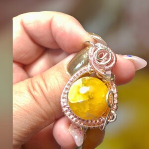 Peut inclure: Un pendentif en pierre jaune avec un enroulement de fil de cuivre. Le pendentif est tenu dans une main.