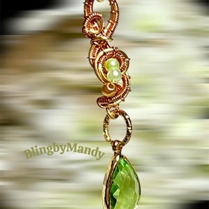 May include: A gold-coloured pendant necklace with a pear-shaped, faceted green gemstone. The necklace has a decorative wire-wrapped design with small green and gold beads. The text "BlingbyMandy" is visible.