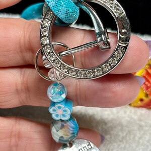May include: A silver charm with rhinestones and a blue mesh charm. The charm has a silver heart-shaped tag with the text "Boots and Bling It's a Cowgirl Thing".