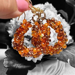 May include: Amber and brown beaded hoop earrings with gold-coloured ear wires. The earrings are handmade and held up by a hand with pink nail polish. The background is a black and white floral arrangement.