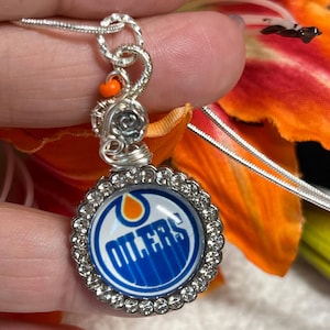 May include: Silver necklace with a round pendant featuring the Edmonton Oilers logo surrounded by clear rhinestones. The logo is blue, orange, and white.