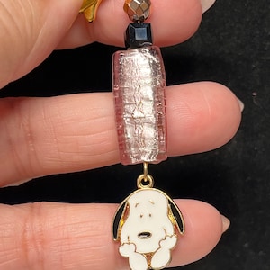 May include: A gold charm featuring a pink and silver glass bead, a black bead, and a gold-coloured metal charm of Snoopy, the Peanuts character, with black ears and a white body.