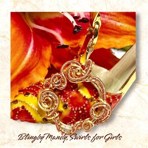 Peut inclure: Un pendentif en fil d'or fait à la main avec des motifs tourbillonnants complexes. Le pendentif est posé sur un fond d'éléments floraux rouges, oranges et jaunes vibrants. Le texte "Blingby Mandy, Swirls for Girls" est visible en bas.