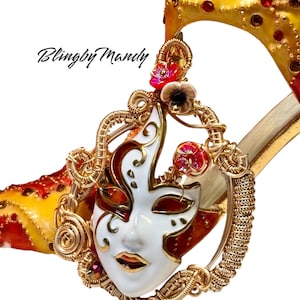 Peut inclure: Gros plan sur une chaussure à talon haut décorative avec un design doré et rouge. La chaussure présente un pendentif en forme de masque blanc et doré avec des accents floraux rouges et roses. Le texte "Blingby Mandy" est visible.