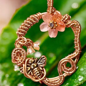 Peut inclure: Pendentif fait à la main de couleur cuivre, orné d'une abeille, de fleurs et d'un motif circulaire. Le pendentif présente une construction délicate en fil de fer, avec une petite fleur rose et un charm en forme d'abeille détaillé. Fond vert.