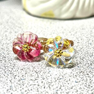 May include: Two flower-shaped rings. One ring features a pink and red flower with a gold centre, while the other has a clear flower with a gold centre. Both rings have gold wire bands.