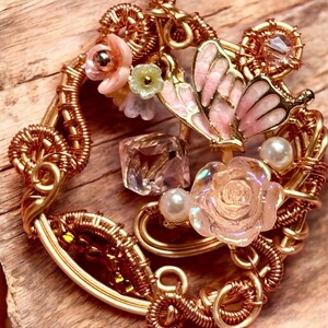 Peut inclure: Un pendentif doré fait à la main avec un papillon aux ailes roses, des fleurs et un cristal transparent. Le pendentif est finement filaire et comprend des perles, créant un design délicat et orné.