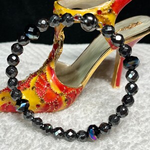 May include: A black faceted bead bracelet with iridescent highlights. The bracelet is displayed in front of a decorative high-heeled shoe with red, yellow, and gold accents. The beads have a geometric cut, reflecting light.