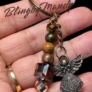 May include: A brown and gold angel charm keychain with a variety of beads, including wood, metal, and glass. The keychain is attached to a silver key ring.