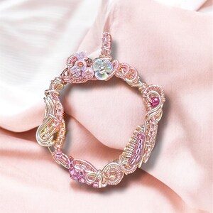 Peut inclure: Un pendentif fait à la main, enroulé de fil métallique, de forme circulaire. Le pendentif présente un mélange de perles et d'accents roses, violets et transparents. Le fil est de couleur cuivre et le pendentif comporte un ornement floral sur le dessus. Le fond est un tissu rose pâle.