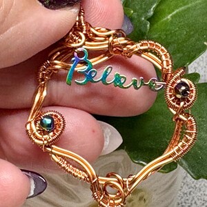 Peut inclure: Pendentif en forme de cœur en fil de cuivre. Le mot "Believe" est écrit en lettres irisées. Le pendentif est orné de petites perles sombres. Le pendentif est tenu par une personne.
