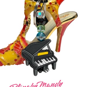 May include: A piano charm keyring with a black and white piano design and beaded accents, set against a decorative high-heeled shoe. The shoe is yellow, red, and gold with rhinestone details. The text "Blingby Mandy" is in pink cursive.