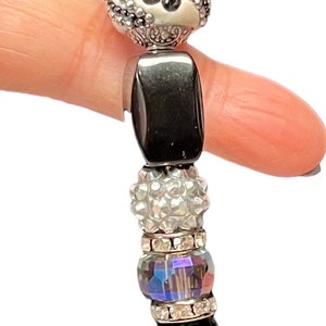May include: A handcrafted beaded pendant with black, silver, and iridescent beads. The pendant includes a faceted black teardrop-shaped bead and a silver wire loop for hanging. The pendant is approximately 10 cm in length.