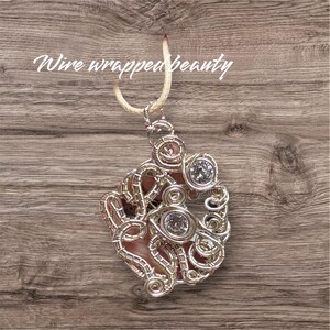 Peut inclure: Pendentif artisanal en fil de fer argenté, orné d'une pierre rose et de deux cristaux ronds et transparents. Le design en fil de fer présente des volutes et des boucles complexes. Le pendentif est suspendu à un cordon beige. Le texte sur l'image est "Wire wrapped beauty".