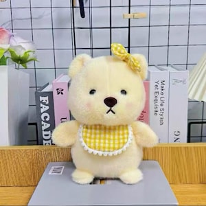 May include: A plush, cream-colored teddy bear with a yellow gingham bib and bow. The bear has a brown nose and black eyes. The bear is standing on a gray surface. The background includes books and decorative items.